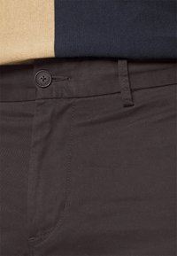 Pantalon marron foncé avec une texture lisse, dotée d'un bouton noir avec un logo, et d'un design à l'avant plat. Partiellement recouvert par un haut beige et marine.
