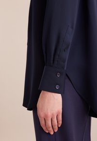 Blusa a maniche lunghe blu navy con una texture liscia, caratterizzata da polsini con bottoni e una vestibilità ampia, mostrata di lato.