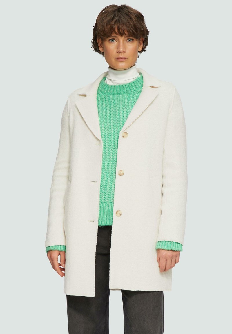 Manteau en laine blanche avec un col cranté, comportant trois boutons et des poches latérales, superposé à un pull en tricot épais vert vif.