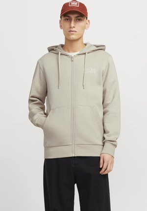 Sudadera con cremallera de color beige claro, con bolsillos delanteros y capucha con cordón, hecha de una tela suave. Presenta un logotipo sutil en el lado izquierdo del pecho.