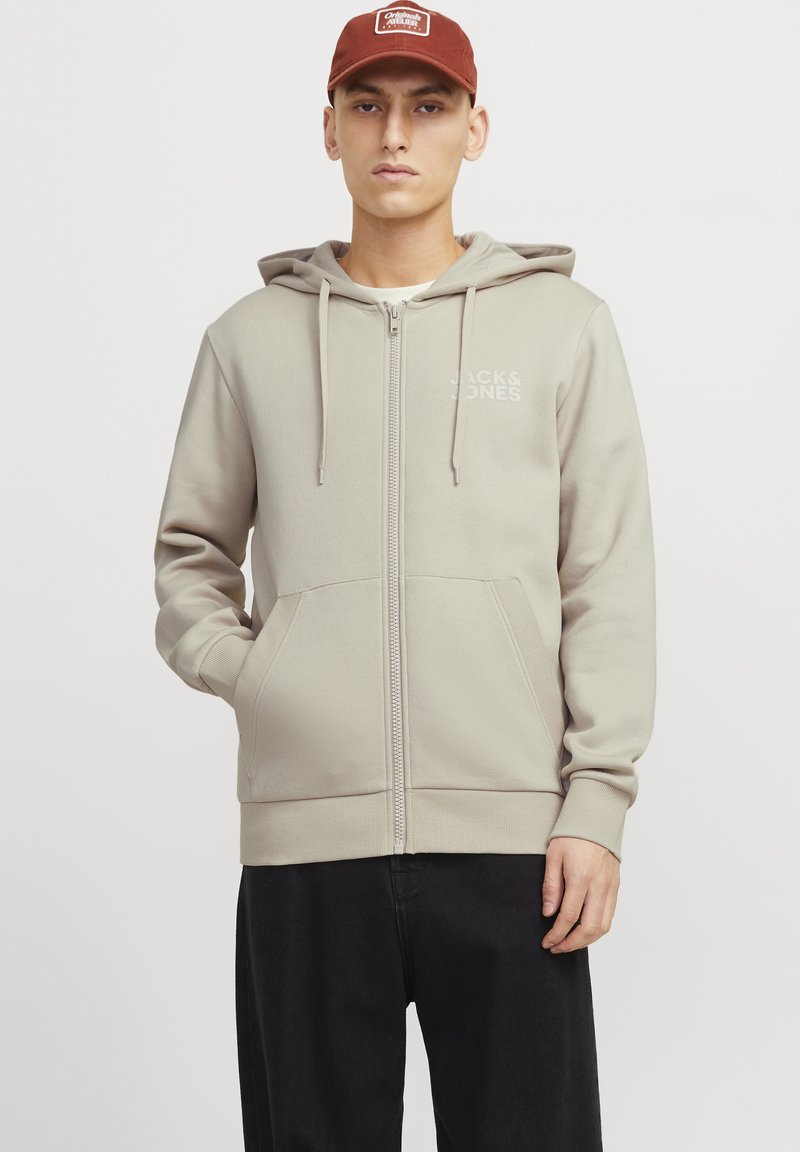 Haut à capuche zippé de couleur beige clair avec poches avant et cordon de serrage, fabriqué en tissu doux. Présente un logo discret sur la poitrine gauche.