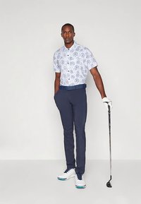 Man draagt een kortarmig overhemd met patroon, marineblauwe broek, witte golfhandschoen en witte golfschoenen, houdt een golfclub vast en staat tegen een effen achtergrond.