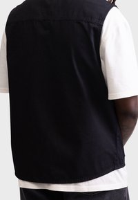Chaleco negro sin mangas de algodón, con una textura suave y dobladillo redondeado, superpuesto sobre una camiseta blanca de mangas cortas. Diseño sencillo.