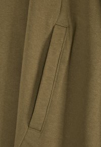 Gros plan sur une poche en tissu vert olive avec des coutures visibles sur un vêtement à texture douce.
