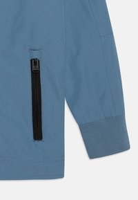 Light blue jacket with a zippered pocket and ribbed cuff. Smooth fabric with stitching detail. Modern design for casual wear.