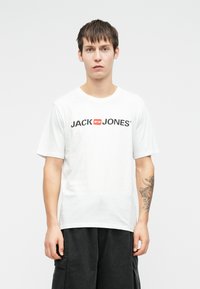 Bílý bavlněný tričko s výrazným černým nápisem "JACK & JONES" s červeným akcentem, v kombinaci s černými cargo šortkami.