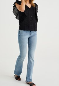 Musta laskostettu paita, jossa on röyhelöpuvut, yhdistettynä vaaleansinisiin leveälahkeisiin farkkuihin ja mustiin kudottuihin sandaaleihin. Napillinen etuosa.