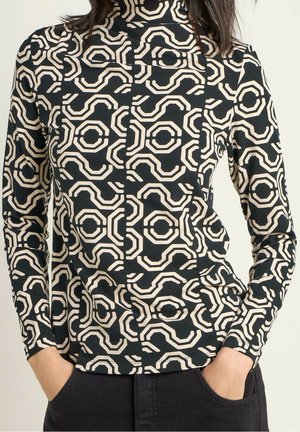 Femme portant un haut à col roulé à manches longues avec motif géométrique noir et beige, les mains dans les poches d’un pantalon noir.