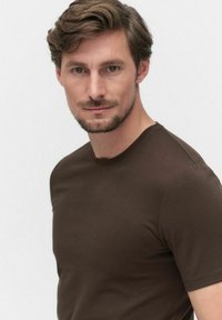 Homme avec les cheveux bruns courts et une barbe, portant un t-shirt à col rond ajusté marron foncé devant un fond clair uni.