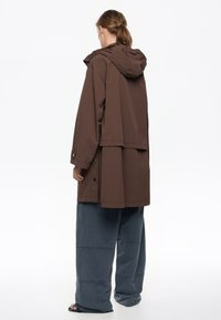 Chaqueta impermeable marrón con capucha, con un corte ligeramente holgado y detalles de botones. Combinada con pantalones anchos grises.