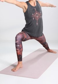 Grijs tanktop met geometrische patronen, gecombineerd met rood-zwart gemêleerde leggings. Model voert een yogahouding uit op een lichtroze mat.