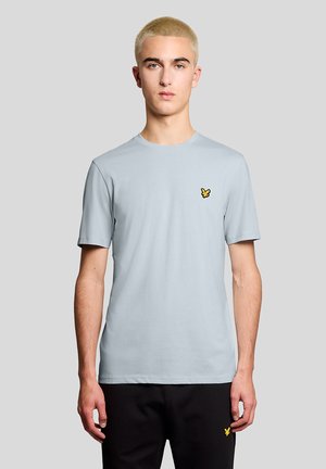 Ung mand med kort blondt hår iført en lyseblå t-shirt og sorte bukser, begge med små gule fuglelogoer på venstre side.