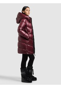 Sieviete, tērpusies garā, spīdīgā bordo krāsas polsterētā mētelī ar kapuci, melnās legingā un melnos ziemas zābakos ar mežģīnēm, stāv sāniski uz pelēka fona.