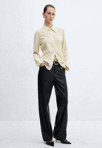 Camicia color crema con colletto grande, abbinata a pantaloni larghi in pelle nera e scarpe nere a punta. Texture lisce, design minimalista.