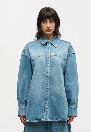 Γυναίκα φοράει υπερμεγέθη μπλε τζιν μπουφάν με γιακά και πέτο στολισμένα με πέρλες, κουμπωμένο μπροστά, στέκεται μπροστά σε απλό φόντο.