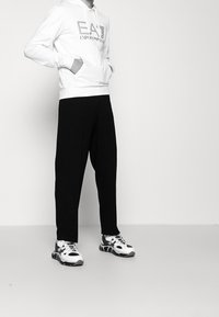 Bílá mikina s kapucí a logem na přední straně, černé volné kalhoty a bílo-černé sportovní boty. Jednoduchý design, pohodlný střih, minimální detaily.