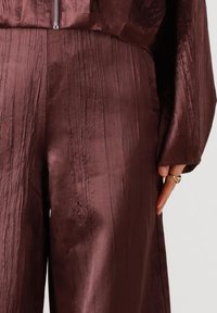 Bordeaux satijnen broek met een getextureerd, geribd oppervlak. Heeft een losse pasvorm, zijzakken en een gladde glans. Geaccentueerd met een gouden ring.