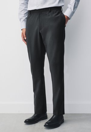 Uomo che indossa pantaloni neri su misura, scarpe in pelle nere e una camicia azzurra a righe con una mano in tasca.