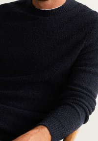 Uomo che indossa un maglione a girocollo testurizzato di un blu scuro, seduto con le braccia incrociate visibili su una sedia di legno.