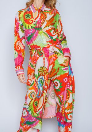Femme portant une longue chemise aux motifs floraux et cachemire multicolores vibrants, associée à un pantalon large assorti avec une ceinture nouée à la taille.