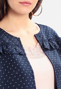 Veste matelassée marine avec des accents à volants et des motifs floraux blancs, portée sur un haut rose clair avec des détails en dentelle au niveau de l'encolure.