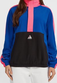 Femme portant une veste à enfiler O'Neill bleue, rose et noire avec un patch logo à l'avant et un pantalon blanc.