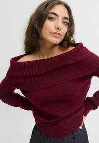 Vīnsarkanā plecu apsedzoša džemperis ar ribotu apkakli un aprocēm. Mīksts adīts tekstūrs, pieguļošs jostasvietā un brīvas piedurknes, valkāts kopā ar melnām biksēm.