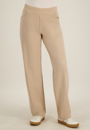 Beige wijde broek met zijzakken, gedragen met witte sneakers met bruine zolen op een effen, neutrale achtergrond.
