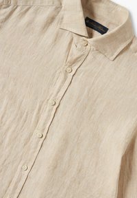 Camicia di lino beige con colletto, chiusura frontale con quattro bottoni e un tessuto leggermente strutturato. Etichetta visibile all'interno del colletto.