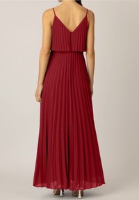 Femme portant une robe rouge sans manches, plissée, longue jusqu'au sol, avec des bretelles fines et un dos en V, debout devant un fond uni.