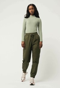 Lichtgroene ribbelige langemouwen top gecombineerd met olijfgroene taps toelopende joggingbroeken. Model draagt crèmekleurige sneakers met donkere accenten. Eenvoudig, casual ontwerp.
