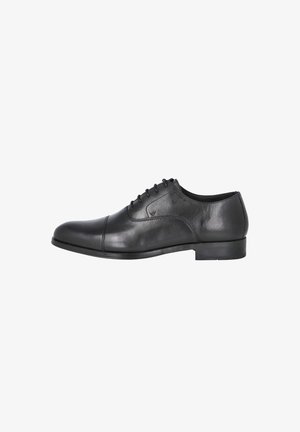 Zapato Oxford de cuero negro con cordones, puntera con costura y tacón bajo, mostrado desde el lado interior sobre un fondo blanco.