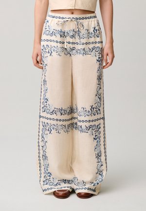 Pantalones sueltos de color crema con estampado floral azul, cintura con cordón y piernas anchas, usados con zapatos marrones.