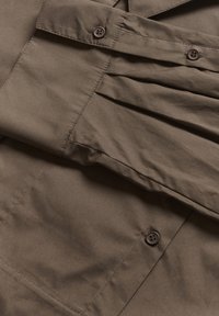 Brown fabric with a smooth texture, featuring pleats and two buttons, creating a structured and functional design.