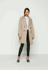 Trench coat bege de duplo peito com botões escuros, combinado com uma blusa branca e calças de imitação de couro pretas. Modelo usa sandálias slides pretas.