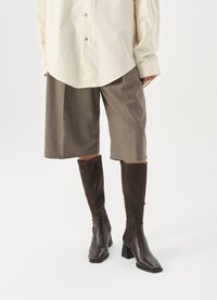 Stivali marroni fino al ginocchio con punta quadrata e tacco a blocco, abbinati a una camicia oversize beige e a pantaloni culottes a quadri marroni.