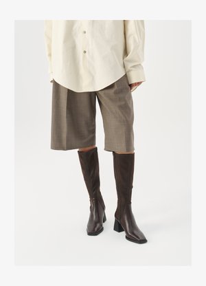 Bottes marron hautes jusqu'au genou avec un bout carré et un talon bloc, associées à une chemise beige surdimensionnée et des culottes à carreaux marron.