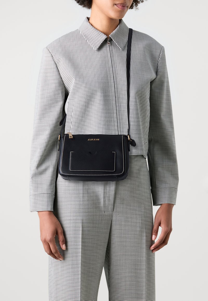 Čierna kožená crossbody taška s bielym prešívaním, s prednou vreckom a zlatým logom. Nastaviteľný popruh a obdĺžnikový tvar.