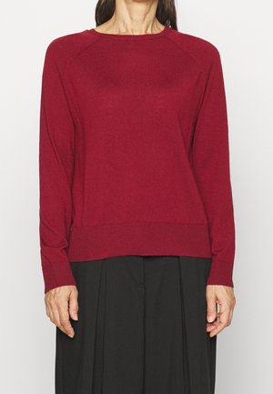 Frau in einem schlichten roten Pullover mit langen Ärmeln, kombiniert mit lockeren schwarzen Hosen, steht vor einem weißen Hintergrund.