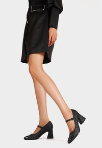 Scarpe con tacco alto nere glitterate con punta arrotondata, accenti di pelle lucida, cinturino alla caviglia e tacco cubano. Superficie texturizzata con lucentezza.