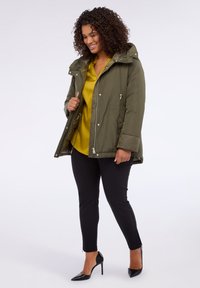 Jaket puffer hijau zaitun dengan penutup kepala, ritsleting depan, dan tali serut. Dipadukan dengan atasan kuning, celana hitam, dan sepatu hak tinggi hitam.