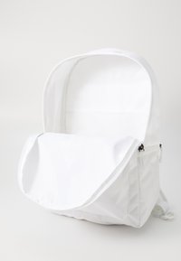 Mochila de tela blanca con un gran compartimento abierto, cierres de cremallera y correas ajustables. La superficie es lisa con un mínimo de detalles.