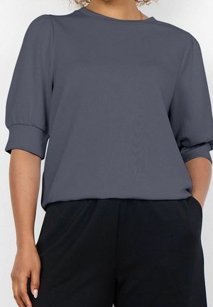Femme portant un haut ample gris foncé à manches trois-quarts et un pantalon noir, debout, une main dans la poche.