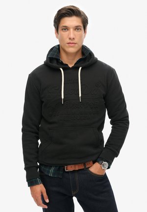 Sudadera negra con un logo texturizado, cordones y bolsillo delantero, superpuesta a una camisa de cuadros azul y verde, combinada con jeans oscuros.