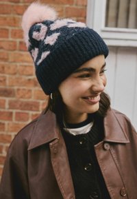 Smiling young woman wearing a navy and pink patterned knit hat with a pompom, brown leather jacket, and black buttoned sweater stands by a brick wall.