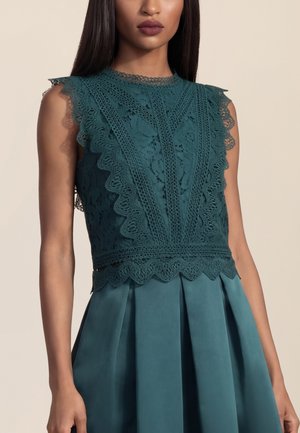 Frau trägt ärmelloses türkisfarbenes Spitzenoberteil mit geschwungenem Saum, kombiniert mit einem passenden plissierten Rock vor beigem Hintergrund.