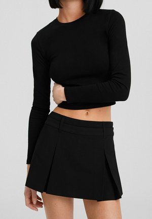 Mujer con top corto de manga larga negro y minifalda plisada negra, posando delante de un fondo liso y claro.