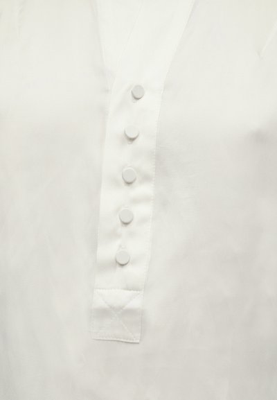 Λευκό μπλουζάκι με απαλό ύφασμα, με λαιμόκοψη σε σχήμα V, έξι στρογγυλά κουμπιά και ραμμένο λοξό κέντημα στο τελείωμα για επιπλέον λεπτομέρεια.