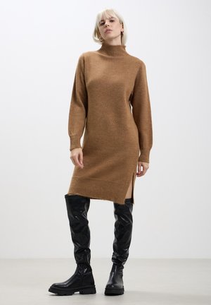Abito di maglia marrone con scollo alto e maniche lunghe, caratterizzato da spacchi laterali e una vestibilità comoda. Abbinato a stivali neri sopra il ginocchio.