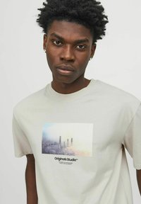Camiseta de algodón gris claro con un estampado gráfico rectangular de un paisaje urbano con niebla, texto negro abajo que dice "Originals Studio™."